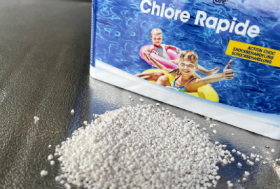 Emballage de granulés chlorés « Chlore Rapide » posé sur une surface sombre, une poignée de granulés blancs est renversée au premier plan. L’emballage montre une photo colorée d’enfants souriants dans une piscine.