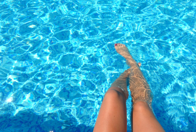 Photo montrant des jambes immergées dans une piscine aux eaux claires et bleu étincelant. La lumière du soleil crée des reflets et des ondulations sur la surface de l’eau, accentuant l’effet cristallin et rafraîchissant. Les jambes croisées, avec des ongles vernis rouges, ajoutent une touche de détente et de sérénité à cette scène estivale. L’image évoque une ambiance de relaxation et de plaisir au bord de l’eau.