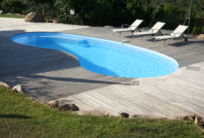 Photo montrant une piscine de forme libre, aux contours arrondis, entourée d'un large deck en bois clair. L'eau de la piscine est d'un bleu éclatant, reflétant la lumière du soleil. À l'arrière-plan, trois chaises longues sont disposées sur le deck, offrant un espace de détente. Environnée de verdure et d'une pelouse bien entretenue à l'avant-plan, cette piscine s'intègre harmonieusement dans un cadre naturel et paisible
