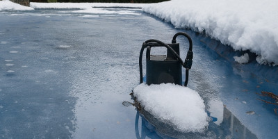 Pompe posée sur une bâche de piscine gelée et recouverte de neige, devant une maison entourée d’arbres en hiver.